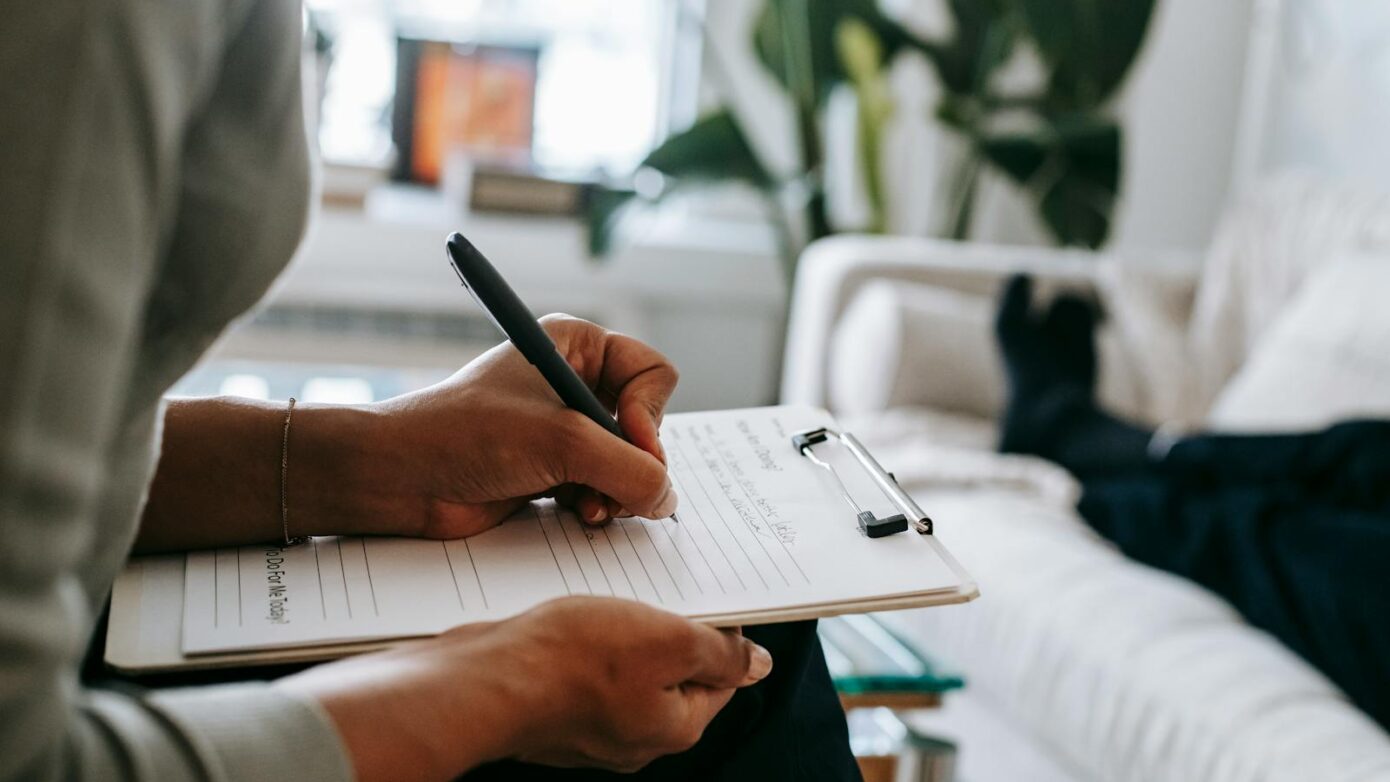 Unrecognizable ethnic female therapist taking notes on clipboard while filling out form during psychological appointment with anonymous client lying on blurred background