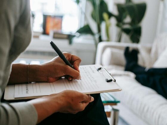 Unrecognizable ethnic female therapist taking notes on clipboard while filling out form during psychological appointment with anonymous client lying on blurred background